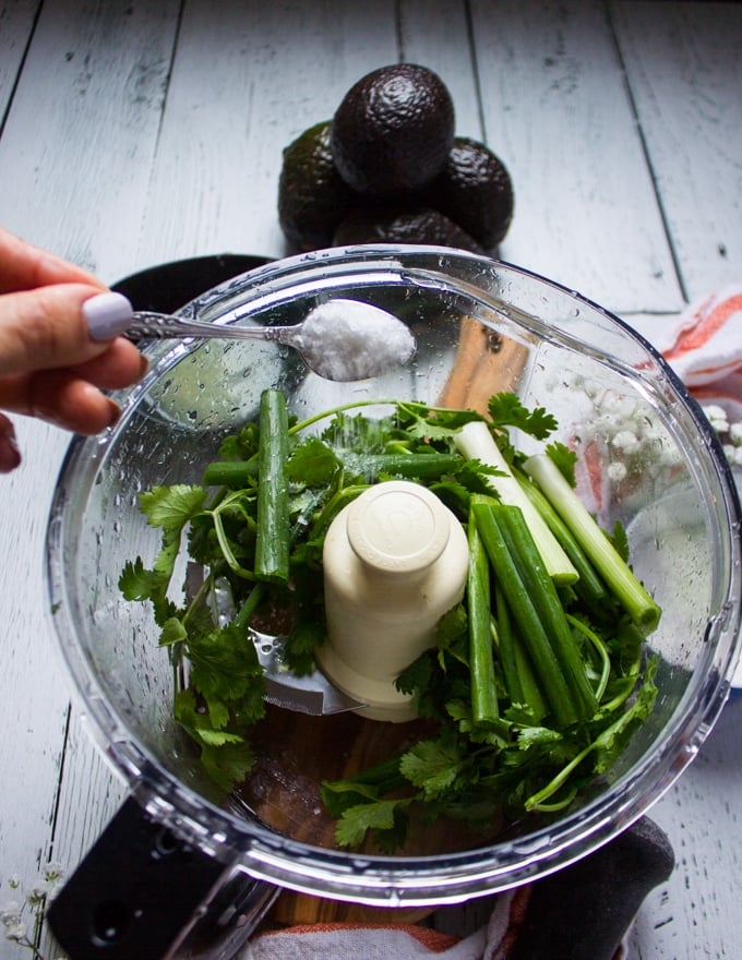 A hand pouring in a spoon of salt in the bowl of a food processor over the cilantro and scallions