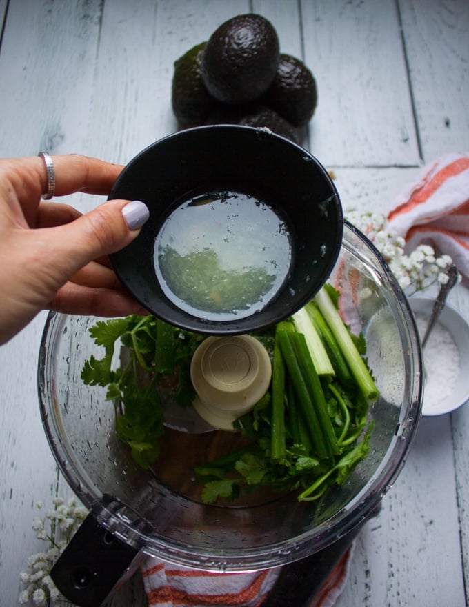 A hand pouring in lime juice from a bowl into the food porcessor with the cilantro and scallions