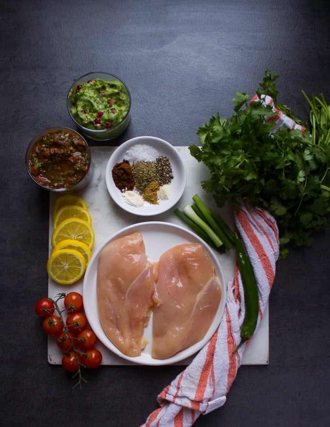 Ingredients for the fajita bowls on a white marble. Chicken breasts on a plate, spice blend on a small plate, salsa, guacamole, cilantro, jalapenos and tomatoes 
