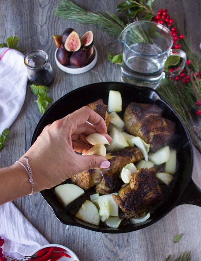 A hand throwing in some whole garlic cloves over the lamb meat and onions in the skillet