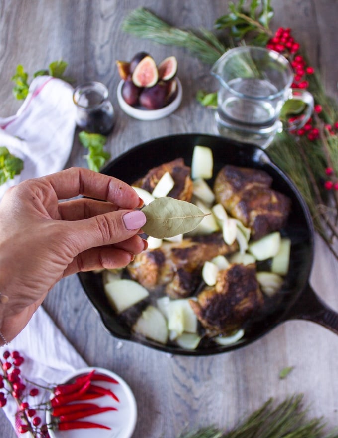 A hand throwing in the bay leaf and cardamom pod over the skillet with lamb meat 