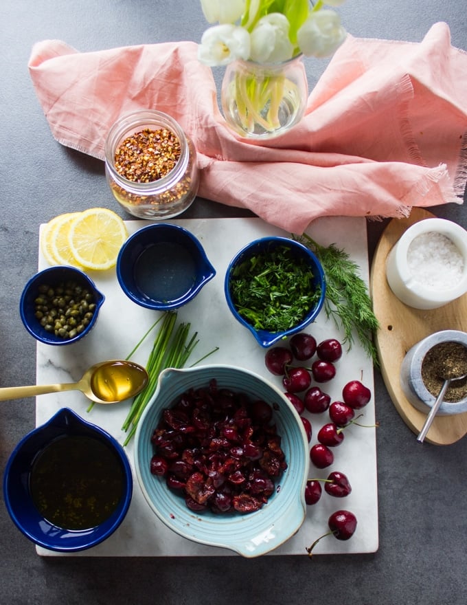 Ingredients for the cherry salsa : cherries in a bowl, salt and pepper, fresh herbs in a bowl, capers in a bowl, olive oil in a bowl, lemon juice in a bowl and a spoon of honey 