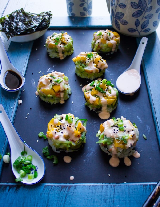 A selection of 8 crab sushi made into sushi sacks over a black board surrounded by a spoon of spicy mayo and a spoon of scallions and soy sauce