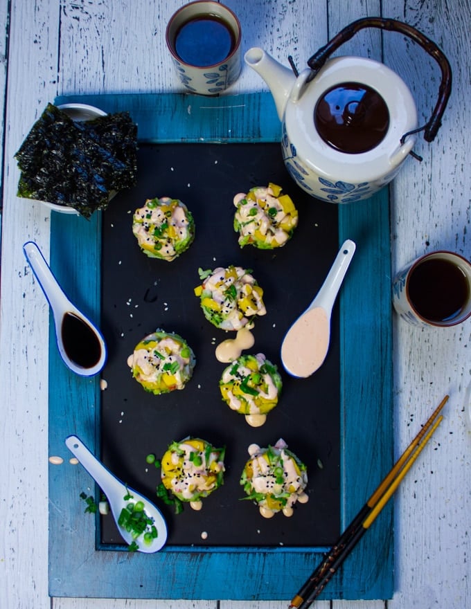 A platter of california rolls made into sushi stacks over a black board surrounded by some Japanese green tea kettle and cups, some chopsticks and spoons of sauces to dip and drizzle. Sauces include spicy mayo and soy sauce