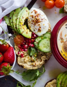 A hummus toast on rye bread with a spread of hummus, a poached egg, some sliced avocados, cucumbers, tomatoes and pomegranate arils