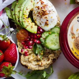 A hummus toast on rye bread with a spread of hummus, a poached egg, some sliced avocados, cucumbers, tomatoes and pomegranate arils