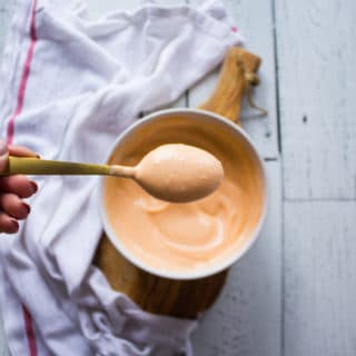 A hand holding a spoonful of spicy mayo over a bowl of spicy mayo recipe surrounded by a tea towel on a white board