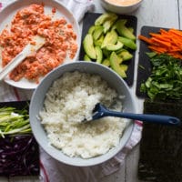 Ingredients for sushi burrito ready to assemble including a bowl of sushi rice, a spicy salmon sushi mixture, fresh vegies chopped for the sushi burrito filling such as sliced avocados, carrots, scallions, purple cabbage and spinach