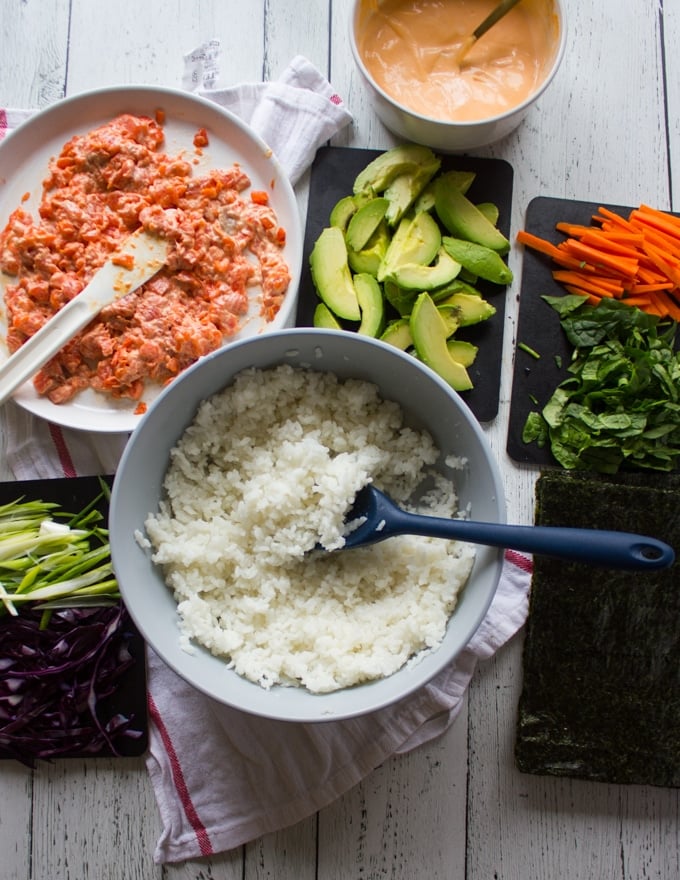 Ingredients for sushi burrito ready to assemble including a bowl of sushi rice, a spicy salmon sushi mixture, fresh vegies chopped for the sushi burrito filling such as sliced avocados, carrots, scallions, purple cabbage and spinach 
