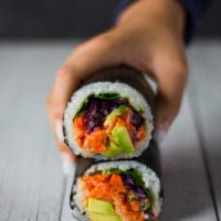 two sushi burrito stacked up on a white board and a hand grabbing the top one. All the filling showing on the inside of the burrito