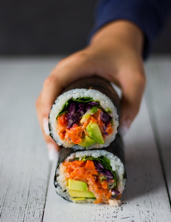 two sushi burrito stacked up on a white board and a hand grabbing the top one. All the filling showing on the inside of the burrito