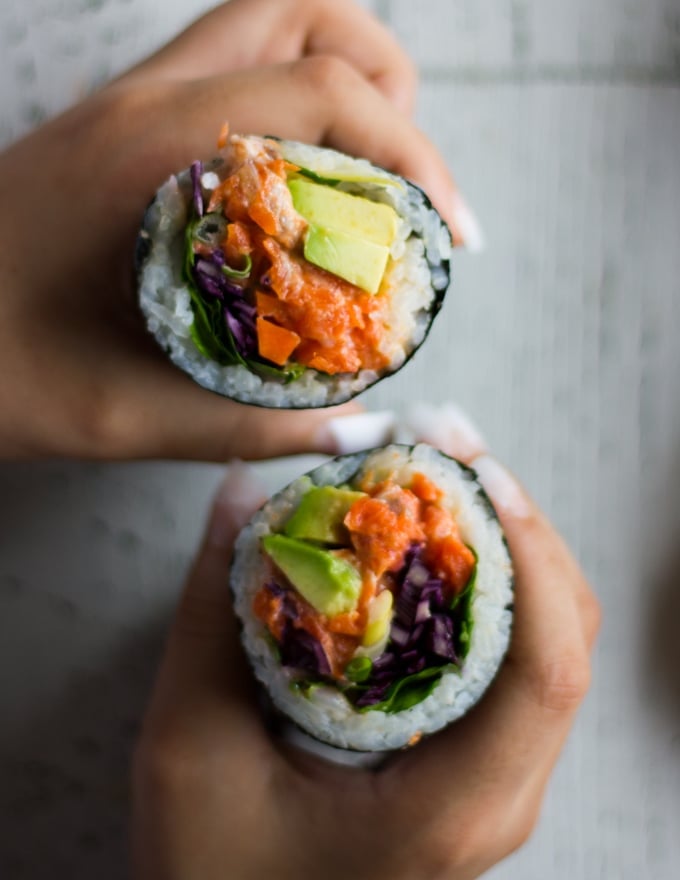 two hands holding off two halves of the sushi burrito pointing up and showing the contrast in colors of the fillings and the sushi rice and all the layers 