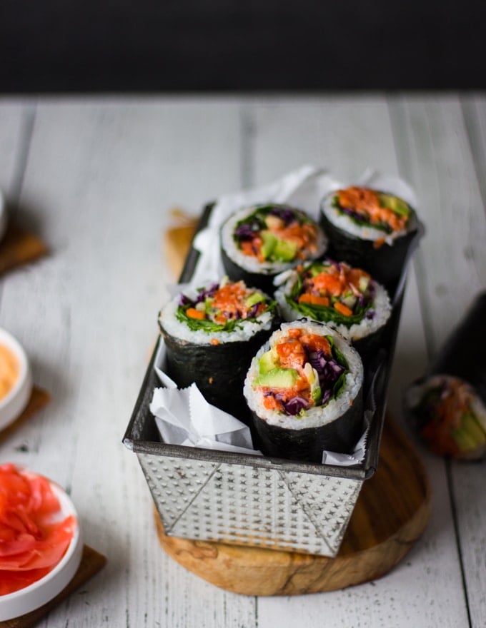 Side view of a rectangle pan lined up with papers and the sushi burritos sliced in half and arranged cut side up in the pan to show the filling and ;ayers . Another sushi burrito next to the pan on the board showing with a plate of spicy mayo