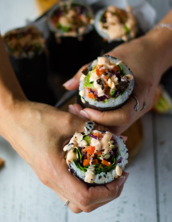two hands holding the sushi burrito finished off with a drizzle of spicy mayo and the sprinkles of crispy salmon skin on top 
