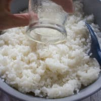 A hand pouring the seasoning from the bowl into the cooled sushi rice to flavor it
