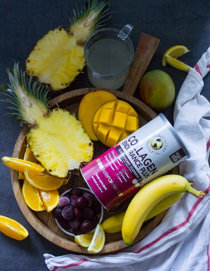 A wooden tray with all the tropical fruits used in the tropical smoothie such as half a pineapple, mangoes, cherries, banana, orange segments and coconut water and a box of collagen to boost the smoothie 
