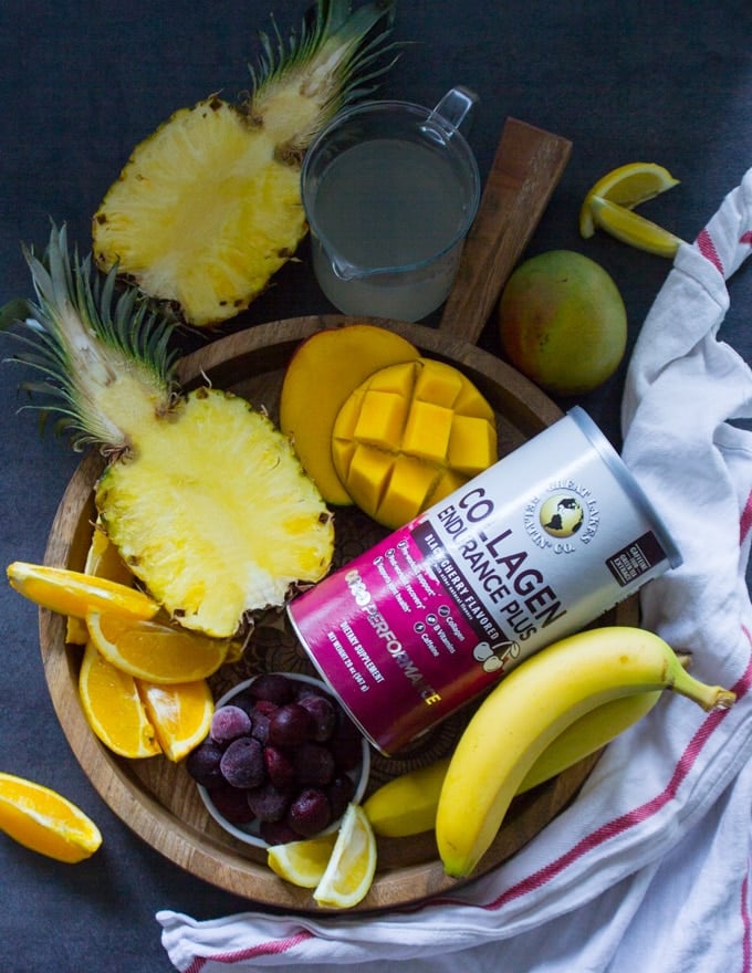 A wooden tray with all the tropical fruits used in the tropical smoothie such as half a pineapple, mangoes, cherries, banana, orange segments and coconut water and a box of collagen to boost the smoothie 