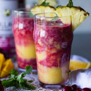 2 cups of tropical smoothies with a cherry smoothie layer and a mango banana smoothie layer in a cup with a pineapple slice on top for garnish