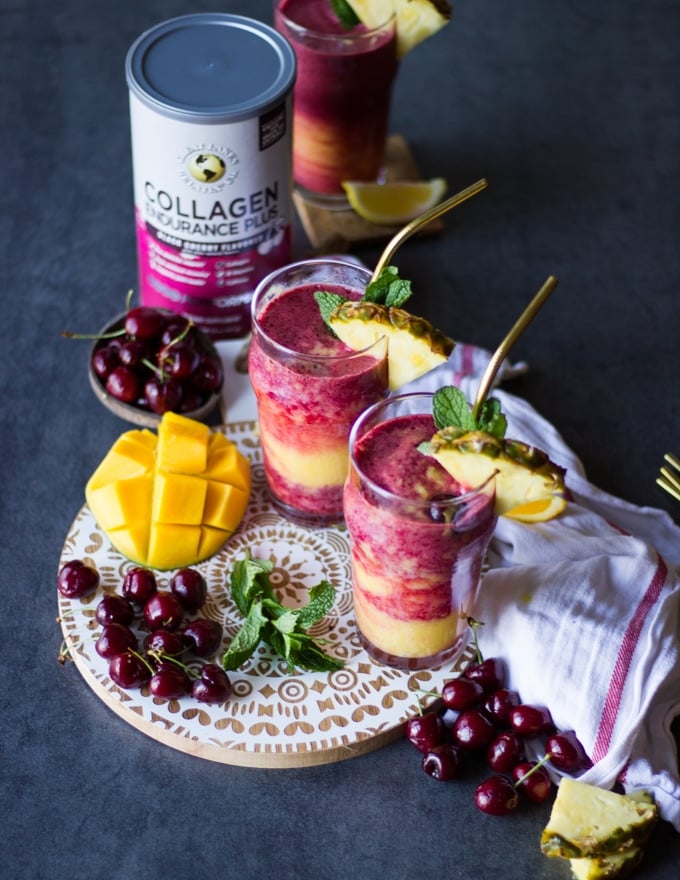 A board with a white wooden top and three cups of tropical smoothies with straws over it, each smoothie garnished with fresh pineapple and surrounded by tropical fruits and a tea towel and the collagen box