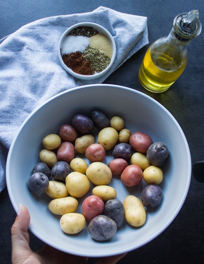 A bowl with baby potatoes raw and a small plate of potato seasoning including, salt, pepper, chilli powder, garlic powder and chilli powder with olive oil