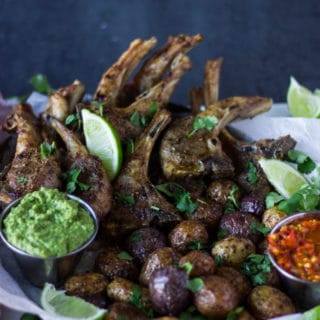 A plate showing crispy air fryer potatoes next to air fryer lamb chops and the poblano sauce in a bowl, sprinkled with cilantro and lime slices