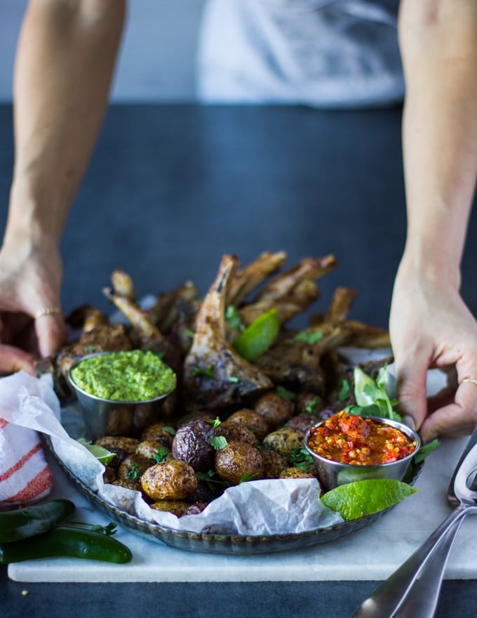 A hand holding the plate of air fryer lamb chops with air fryer potatoes, the poblano sauce in a bowl and the hot pepper sauce in another bowl