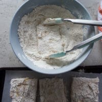 The fish fillets being dredged once again in the flour bowl and a tong is used to coat it