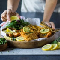 A hand holing the fish fry plate with french fries and the other hand squeezing lemon juice over the fried fish