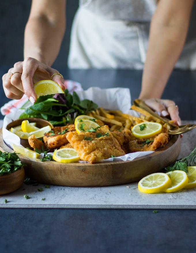 A hand holing the fish fry plate with french fries and the other hand squeezing lemon juice over the fried fish