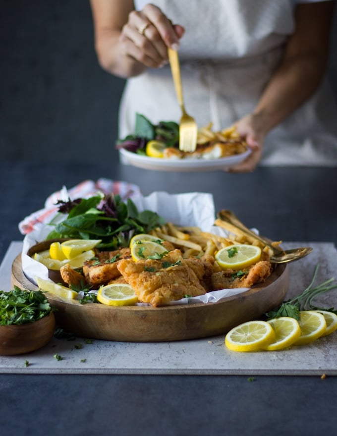 A huge plate of fish fry, french fries, greens with lemon slices and at the back someone holding a plate of fried fish and eating it with a fork