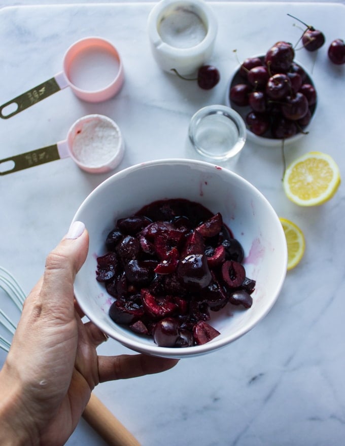 A hand holding a bowl of frozen cherries to make cherry pie filling with the remaining ingredients in a white marble below. 