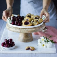 A stand with a white plate filled with mini cherry pie and some fresh cherries on the side, a pink napkin and white flowers. Two hands holding the plate.