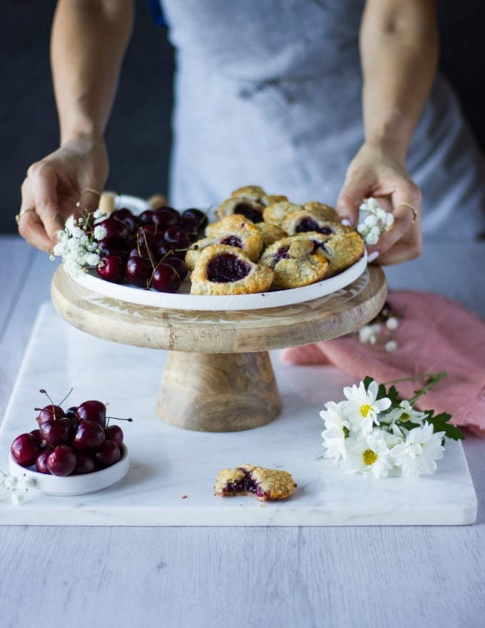 A stand with a white plate filled with mini cherry pie and some fresh cherries on the side, a pink napkin and white flowers. Two hands holding the plate.