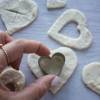 A hand cuting out smaller heart shapes on the tops of the pie crusts for cherry pie