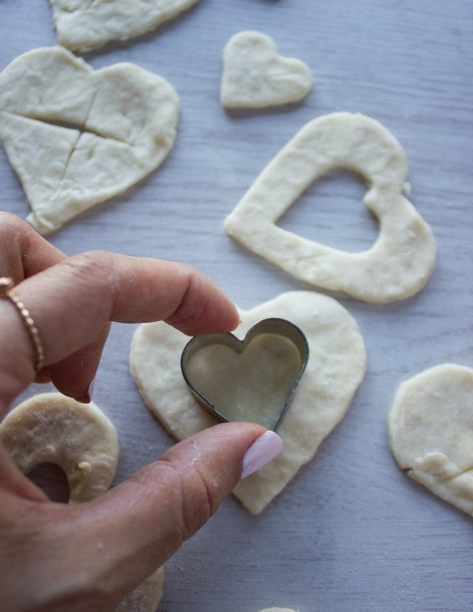 A hand cuting out smaller heart shapes on the tops of the pie crusts for cherry pie