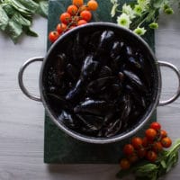 A colander of fresh mussels on a green marble surrounded by basil and fresh and tomatoes