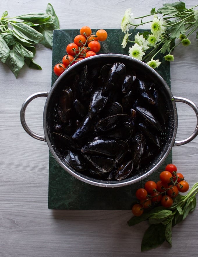 A colander of fresh mussels on a green marble surrounded by basil and fresh and tomatoes