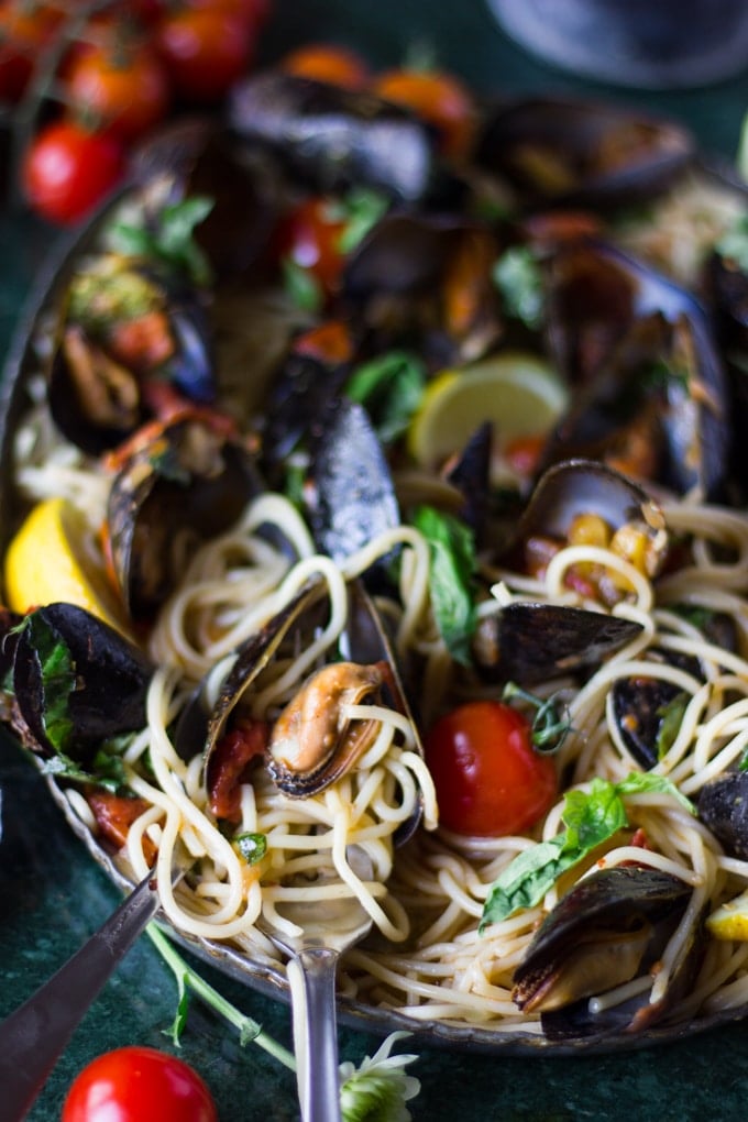 Close up of a spoon in a bowl of mussels pasta showing the tomato marinara sauce, the cooked mussels, the fresh basil and lemon wedges