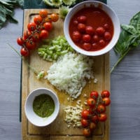 Ingredients for mussels marinara including chopped onions, celery, garlic, basil pesto and canned tomatoes on a cutting board