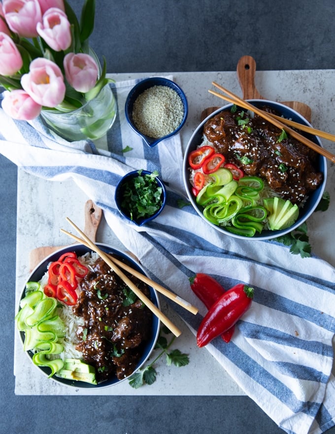 2 ready made bowls of beef tips and rice with a blue towel and some pink tulips, a bowl of cilantro and sesame seeds, some avocado slices and chopsticks to serve