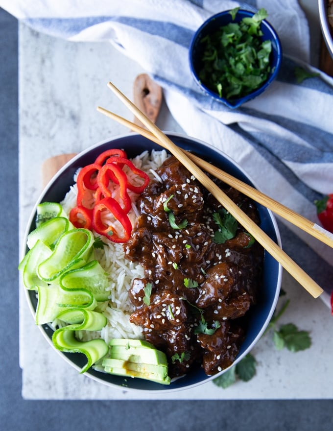 A bowl of beef tips and rice with chopsticks, cucumber rounds red bell peppers and cilantro in a small bowl with a blue towel on the side