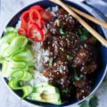 A bowl of beef tips and rice with chopsticks, cucumber rounds red bell peppers and cilantro