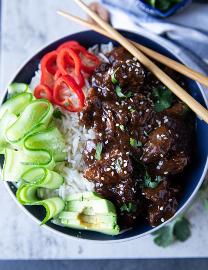 A bowl of beef tips and rice with chopsticks, cucumber rounds red bell peppers and cilantro