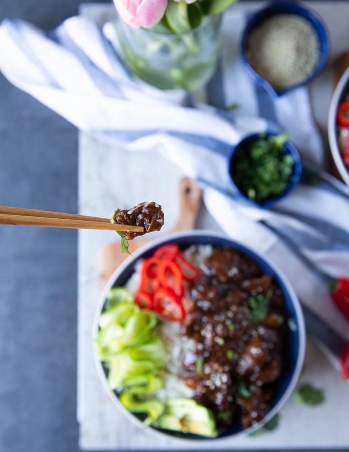 One chop stick holding a beef tip over a bowl of beef tips and rice showing the texture and sauce of the beef tips 