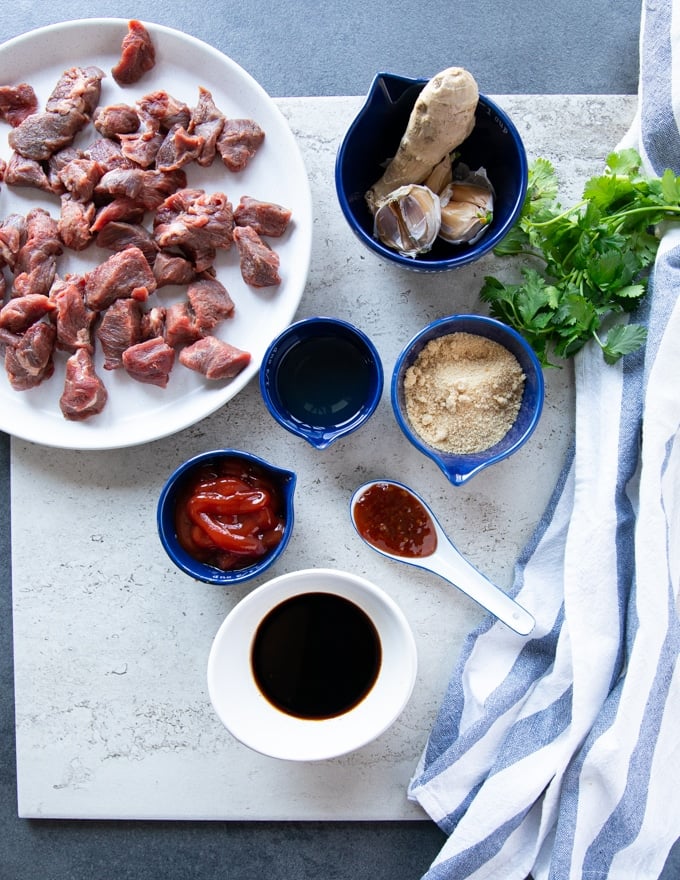 Ingredients for beef tips recipe: beef tips on a plate, fresh ginger and fresh garlic, brown sugar, ketchup, soy sauce, rice vinegar, chilli sauce, cilantro