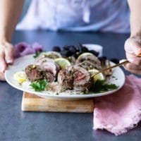Two hands holding a plate of roast boneless leg of lamb over rice, sliced thinly and one hand serving the lamb using a spoon