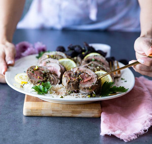 Two hands holding a plate of roast boneless leg of lamb over rice, sliced thinly and one hand serving the lamb using a spoon