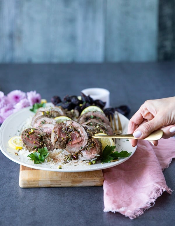A hand holding a spoon serving one piece of the sliced roasted leg of lamb showing the lamb