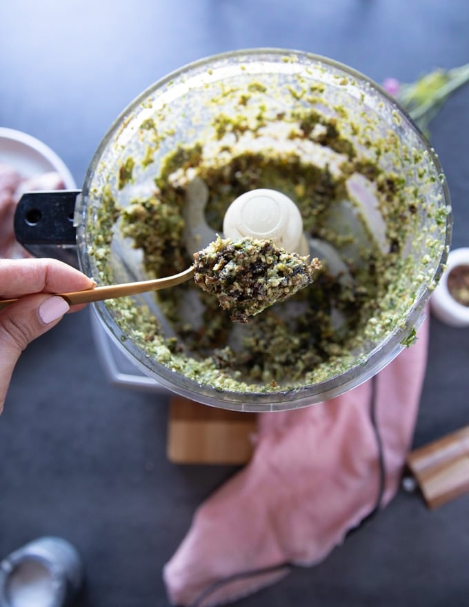 A hand holding a spoon over the food processor showing the filling for the leg of lamb