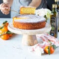 A hand holding a slice of olive oil cake over the serving olate with some clementine oranges and a tea towel around it